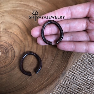 May include: Two dark brown wooden hoop earrings. The earrings are made of wood with a natural grain pattern. The earrings are shown on a hand, with the larger earring on the palm and the smaller earring on the fingers.