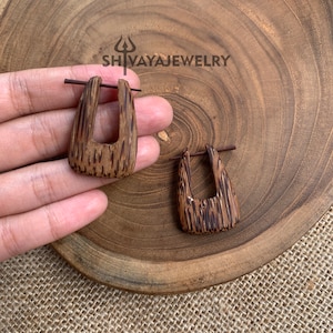 May include: A pair of brown wooden earrings with a rectangular shape and a cutout in the centre. The earrings are made of natural wood and have a unique grain pattern. The earrings are shown on a wooden surface.