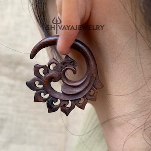May include: A dark brown wooden ear gauge with a carved floral design. The gauge is shaped like a spiral and has a decorative, intricate pattern.