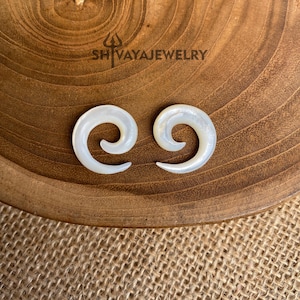 Peut inclure: Deux jauges d'oreille en spirale de coquille blanche sur une surface en bois. Les jauges sont faites de coquille naturelle et ont une finition lisse et polie.