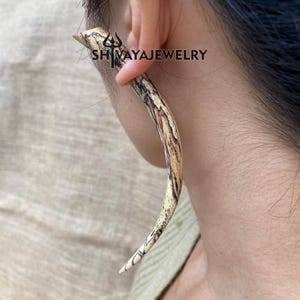 May include: A curved, wooden earring with a natural, marbled pattern of light brown and dark brown. The earring is worn in the ear. The text "SHIVAYAJEWELRY" is visible on the ear. The earring is a unique piece of jewelry.