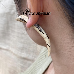May include: A curved, horn-shaped earring with a natural wood grain pattern of cream and black. The earring is inserted into an ear, with the text "SH VAYAJEWELRY" visible.
