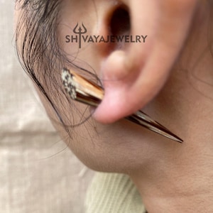May include: Close-up of a brown and white earring in the shape of a spike, worn in an ear piercing. The earring has a pointed end and a textured surface. The brand name "SH VAYAJEWELRY" is visible.