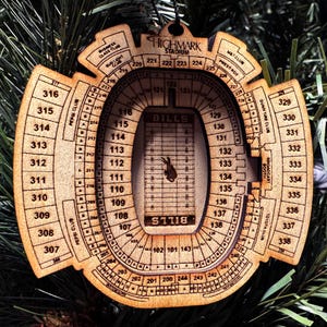 May include: A wooden ornament shaped like Highmark Stadium, home of the Bills. The ornament is light brown and features detailed cutouts of the stadium's seating sections, labeled with numbers. The word "BILLS" is visible on the field. A perfect gift for any Bills fan.