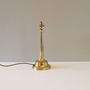 May include: A brass lamp base with a fluted column and a circular base. The lamp has a standard bulb holder and a brown electrical cable. The lamp is on a white surface against a grey background.