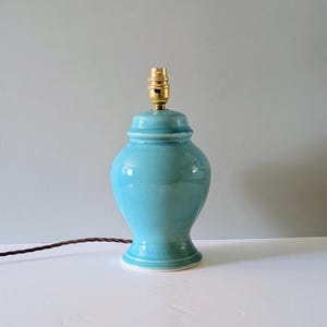 May include: A turquoise ceramic table lamp base with a glossy, crackled glaze finish. The lamp features a classic urn-shaped body with a flared base and a metallic gold-toned light bulb socket at the top, accompanied by a brown twisted fabric cord.