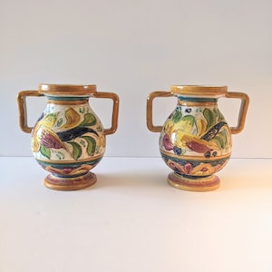 May include: Two small ceramic vases with handles. The vases are decorated with colourful floral patterns and have a yellow, brown, and white base.
