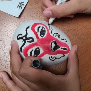 May include: A person is shown drawing on a white, oval-shaped object with a red and black face design. A paper with the same design and Japanese text is next to it. The person is using a black marker to add details to the face.