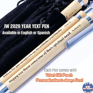 May include: A close-up of several pens with silver accents and light-colored wooden barrels. The pens have engraved text in English and Spanish. The image also shows a black velvet pouch. The text on the pens includes "JW 2026 YEAR YEXT PEN" and "Happy are those conscious of their spiritual need."