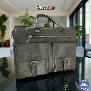 A dark brown vegan leather briefcase with two front buckled pockets, a top zipper, and two handles, rests on a black reflective surface.
