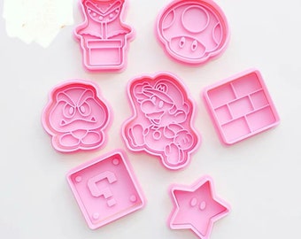 May include: A collection of seven pink plastic cookie cutters in various shapes, including a mushroom, a star, a question mark, and a character resembling Mario. The cutters are designed for creating themed cookies.
