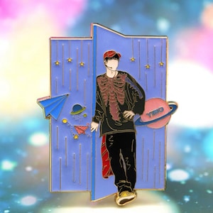 May include: A gold-toned enamel pin featuring a cartoon illustration of a person wearing a red hat, black shirt, and black pants. The person is standing in front of a blue door with stars and a paper airplane. The planet next to the person says "Yibo".