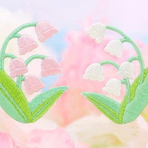Op de afbeelding: Twee geborduurde pleisters met lelietjes-van-dalen. Eén pleister heeft roze bloemen en de andere heeft witte bloemen. Beide pleisters hebben groene bladeren.