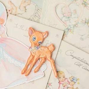 May include: A light brown embroidered fawn with a floral wreath around its neck. The fawn is on a white background with pink and blue accents.