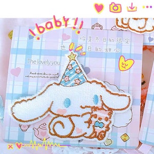 Puede incluir: Un parche bordado de felpa blanca con un perro de dibujos animados que lleva un gorro de fiesta a rayas azules y blancas. El perro sostiene un perro de felpa más pequeño. El parche está sobre un fondo a cuadros rosa y azul con el texto "The lovely you" y "你是冬日的限定 也反日的甜心" en caracteres chinos.