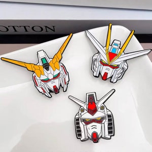 May include: Three enamel pins featuring the heads of different robots from the anime series "Gundam". The pins are white with red, yellow, and blue accents.