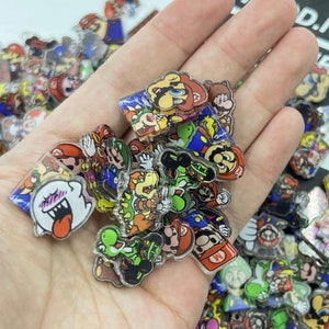 May include: A collection of colorful, cartoon character charms featuring characters from the Super Mario Bros. video game franchise. The charms are made of clear plastic with printed images of Mario, Luigi, Peach, Toad, Bowser, Yoshi, and other characters.
