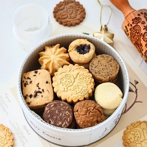 Può includere: Una scatola rotonda bianca piena di diversi tipi di biscotti. I biscotti sono decorati con diversi motivi e disegni. La scatola ha le parole "more cookie" stampate.