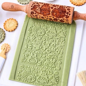 May include: A wooden rolling pin with a carved design of a cartoon character. The rolling pin is on top of green dough with the same design imprinted on it. The dough is on a white surface. There are also cookies with the same design on the surface.