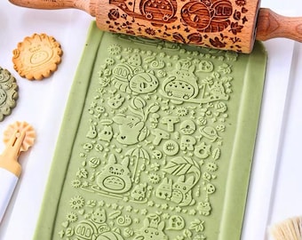 Puede incluir: Un rodillo de madera con un diseño tallado de un personaje de dibujos animados. El rodillo está encima de una masa verde con el mismo diseño estampado en ella. La masa está sobre una superficie blanca. También hay galletas con el mismo diseño en la superficie.