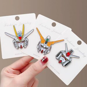 May include: Three white card stock tags with the text "Fashion Jewelry" printed on them. Each tag features a different anime robot head design in enamel pin form. The pins are white, red, yellow, and blue.