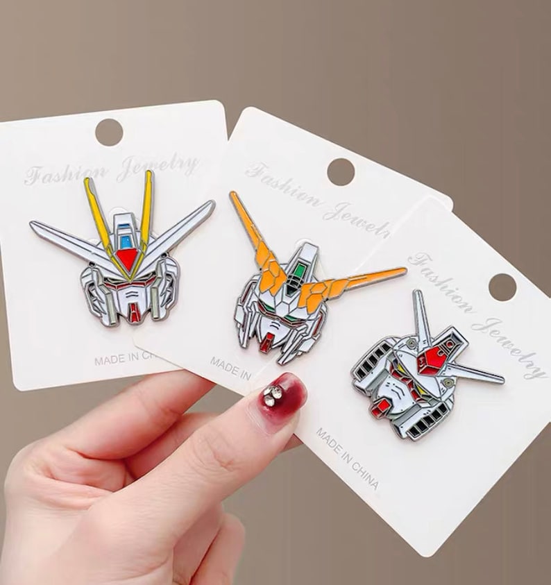 May include: Three white card stock tags with the text "Fashion Jewelry" printed on them. Each tag features a different anime robot head design in enamel pin form. The pins are white, red, yellow, and blue.