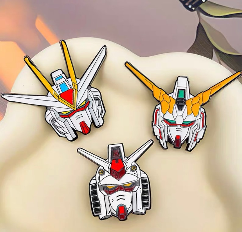 May include: Three enamel pins featuring the heads of different robots from the anime series "Gundam". The pins are white, red, yellow, and blue.