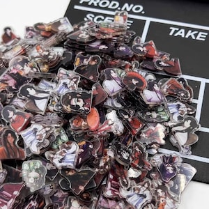 Może przedstawiać: Duża sterta przezroczystych plastikowych zawieszek z postaciami anime. Zawieszki są w różnych pozach i wyrażeniach, niektóre trzymają przedmioty, takie jak miecze i kwiaty. Zawieszki znajdują się na czarnym tle z widocznymi napisami "PROD. NO." i "TAKE".