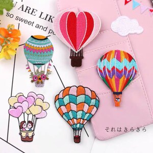 Peut inclure: Assortiment de patchs brodés colorés à repasser représentant des montgolfières de formes et de motifs variés. Les patchs comprennent une montgolfière en forme de cœur, une montgolfière à motif floral, une montgolfière à rayures et une montgolfière à carreaux. Il y a aussi un patch en forme de nuage et un patch avec un couple dans une montgolfière.