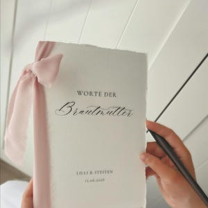 Peut inclure: Une carte blanche avec le texte "WORTE DER Brautmutter" en écriture élégante, attachée avec un ruban rose. La carte comprend également les noms "LILLI & STEFFEN" et la date "12.06.2026".