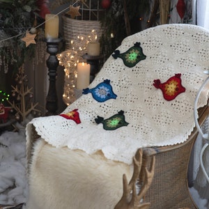 Puede incluir: Una manta de ganchillo blanca con dise&ntilde;os de p&aacute;jaros coloridos. La manta est&aacute; drapeada sobre una silla de mimbre con un coj&iacute;n de piel sint&eacute;tica y una figura de ciervo dorada.