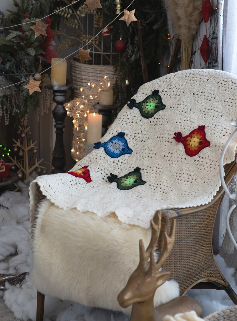 Puede incluir: Una manta de ganchillo blanca con dise&ntilde;os de p&aacute;jaros coloridos. La manta est&aacute; drapeada sobre una silla de mimbre con un coj&iacute;n de piel sint&eacute;tica y una figura de ciervo dorada.