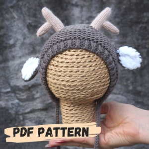 May include: A brown knitted baby hat with deer antlers and ears. The hat has a white pom-pom on each ear. The hat is on a brown mannequin head. The text "PDF PATTERN" is written on a white and orange banner.