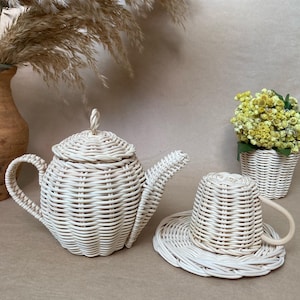 Puede incluir: Un juego de té de mimbre blanco con una tetera, una taza y un platillo. El juego está hecho de mimbre tejido y tiene un aspecto natural y rústico.