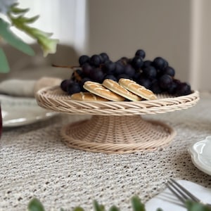 Puede incluir: Un soporte para pasteles de mimbre tejido sostiene un racimo de uvas moradas oscuras y varias galletas pequeñas y oblongas. El soporte es redondo con una base de pedestal y se asienta sobre un mantel beige texturizado. Se ven un tenedor y un plato.