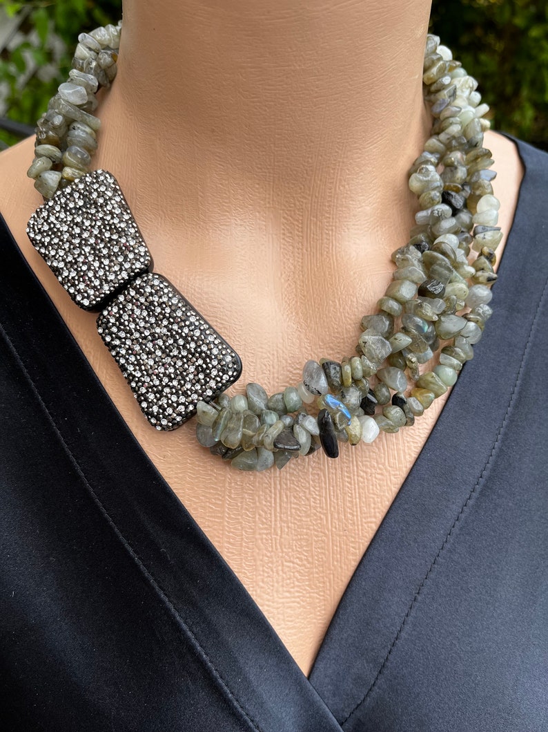 May include: A multi-strand necklace made of grey and black stones with a large, black, crystal-encrusted bow accent.