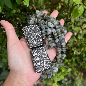 May include: Two silver-toned rectangular clasps with a sparkly, crystal-like finish. The clasps are held in a hand, and behind them is a strand of grey and green gemstone beads.
