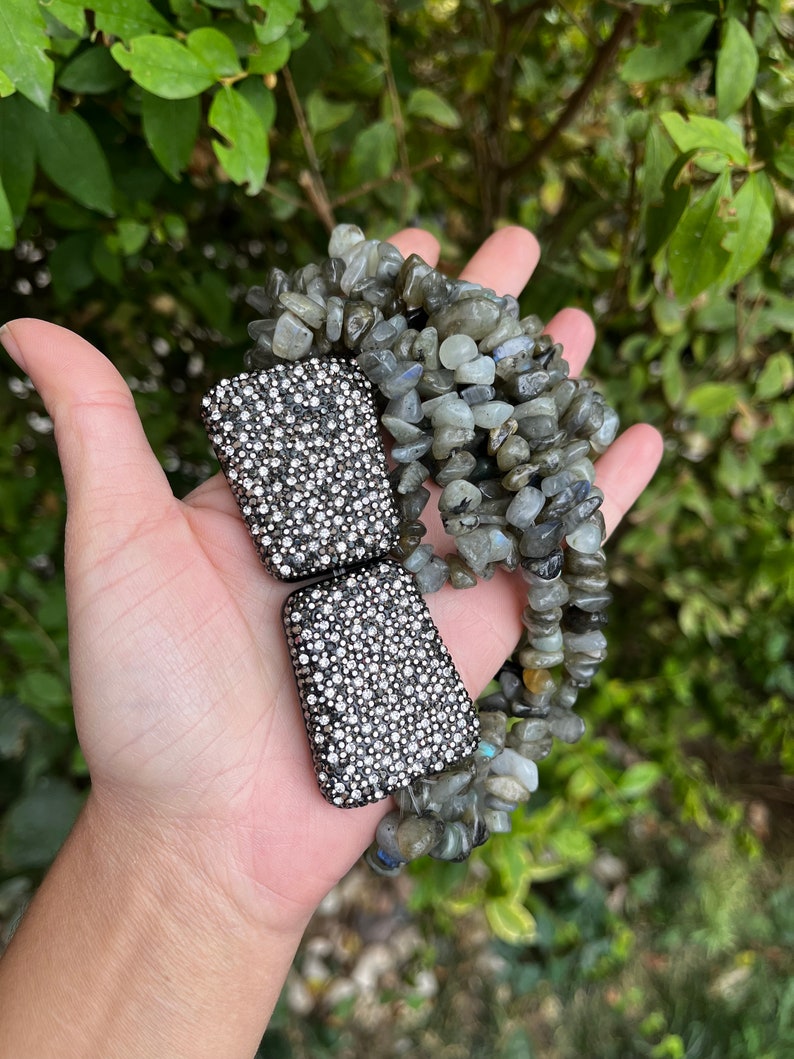 May include: Two silver-toned rectangular clasps with a sparkly, crystal-like finish. The clasps are held in a hand, and behind them is a strand of grey and green gemstone beads.