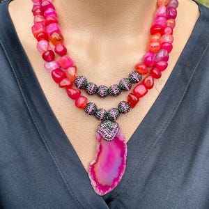 Puede incluir: Un collar de varias capas con cuentas de ágata rosa y roja y un gran colgante de ágata rosa con detalles negros y rosas. El collar está sobre un fondo negro.