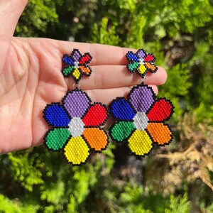 Puede incluir: Un par de pendientes de flores con cuentas. Cada pendiente presenta una flor grande y multicolor con pétalos en tonos de azul, morado, rojo, naranja, amarillo y verde, delineados en negro. Los pendientes están sostenidos por un gancho plateado.