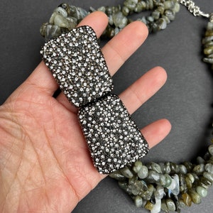 May include: A multi-strand necklace made of green and gray stones with a silver clasp. The clasp is decorated with a large, rectangular, silver-toned piece that is covered in small, clear rhinestones.