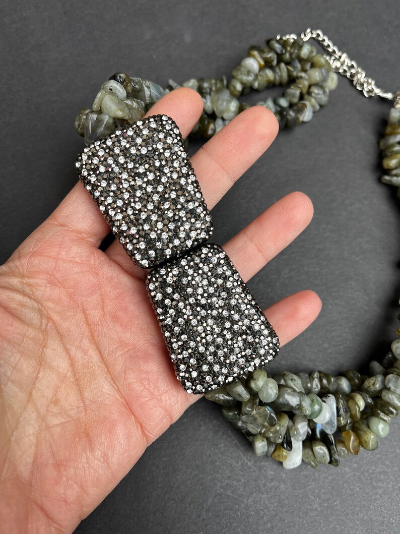 May include: A multi-strand necklace made of green and gray stones with a silver clasp. The clasp is decorated with a large, rectangular, silver-toned piece that is covered in small, clear rhinestones.