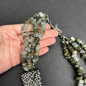May include: A multi-strand necklace made of green and gray stones with a large, black, rhinestone-encrusted pendant.