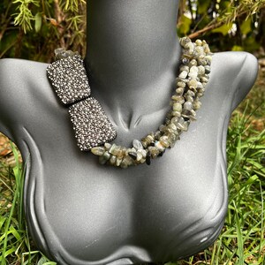 May include: A multi-strand necklace made of grey and black stones. The necklace has a silver-toned clasp and is displayed on a grey mannequin.