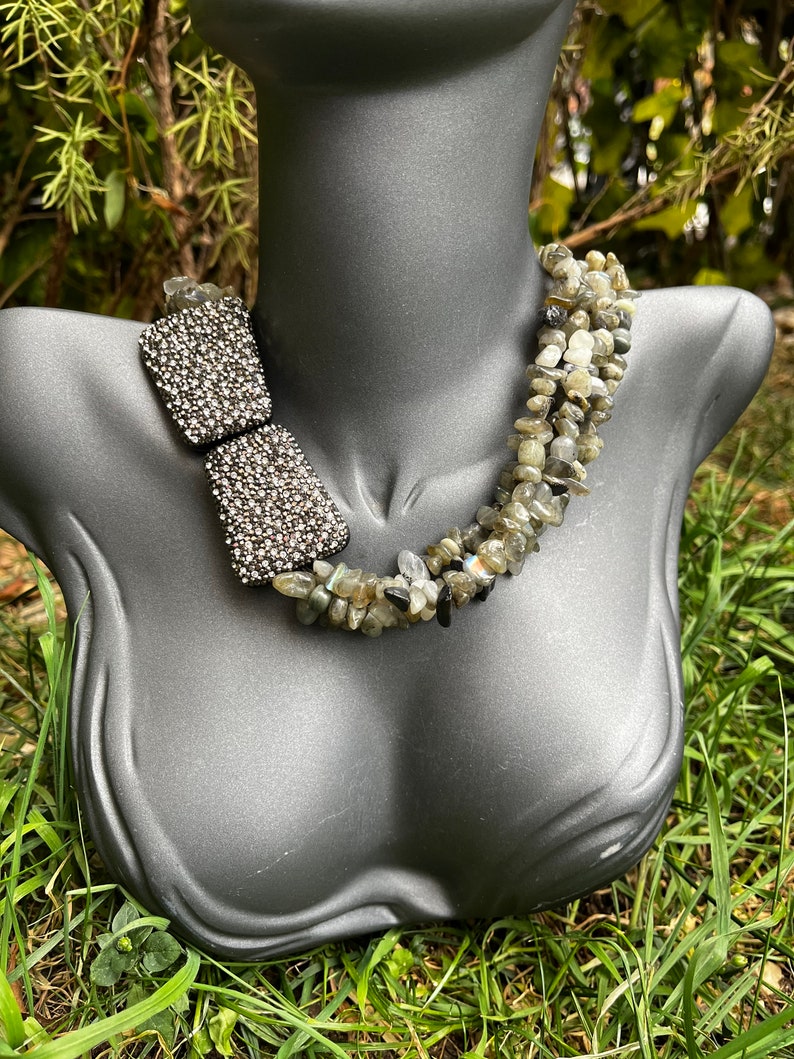 May include: A multi-strand necklace made of grey and black stones. The necklace has a silver-toned clasp and is displayed on a grey mannequin.