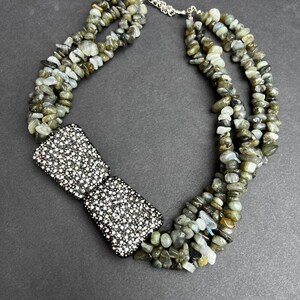 May include: A multi-strand necklace made of grey and green gemstone beads with a large, square, silver-toned rhinestone bow pendant.