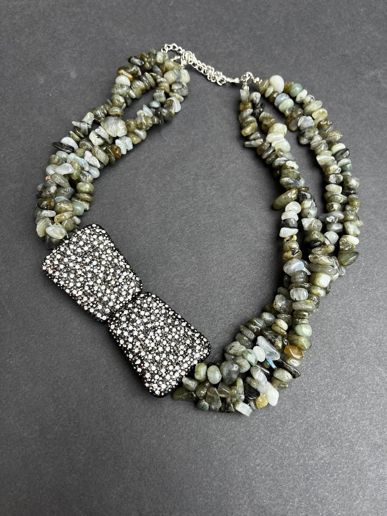 May include: A multi-strand necklace made of grey and green gemstone beads with a large, square, silver-toned rhinestone bow pendant.