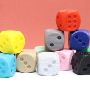 Puede incluir: Un conjunto de dados coloridos en varios colores, incluyendo rojo, azul, amarillo y gris. Cada dado tiene un número diferente de puntos, dispuestos en la configuración estándar. Los dados están apilados unos sobre otros.