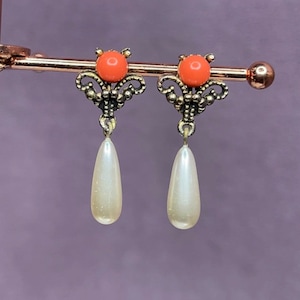 Puede incluir: Un par de pendientes de gota de tono dorado con una cuenta de color coral en la parte superior y una gota nacarada blanca.