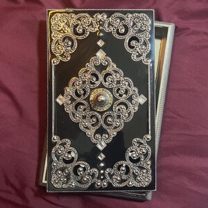 May include: A black compact mirror with ornate silver filigree and sparkling crystal accents. The compact has a rectangular shape and a hinged opening, revealing a mirror inside. The design is symmetrical and elegant.